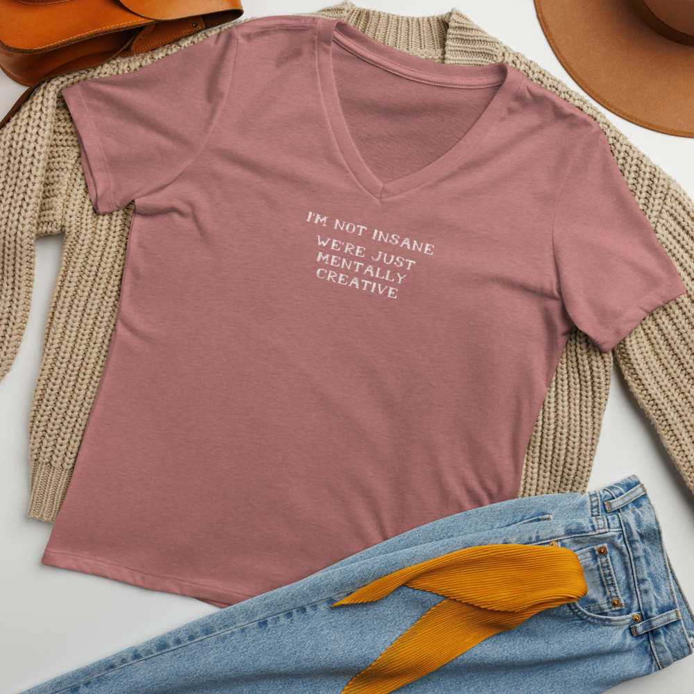 "I’m not insane   We’re just mentally creative" printed on a heather mauve women's relaxed v-neck t-shirt laying on a flat surface amongst other clothes