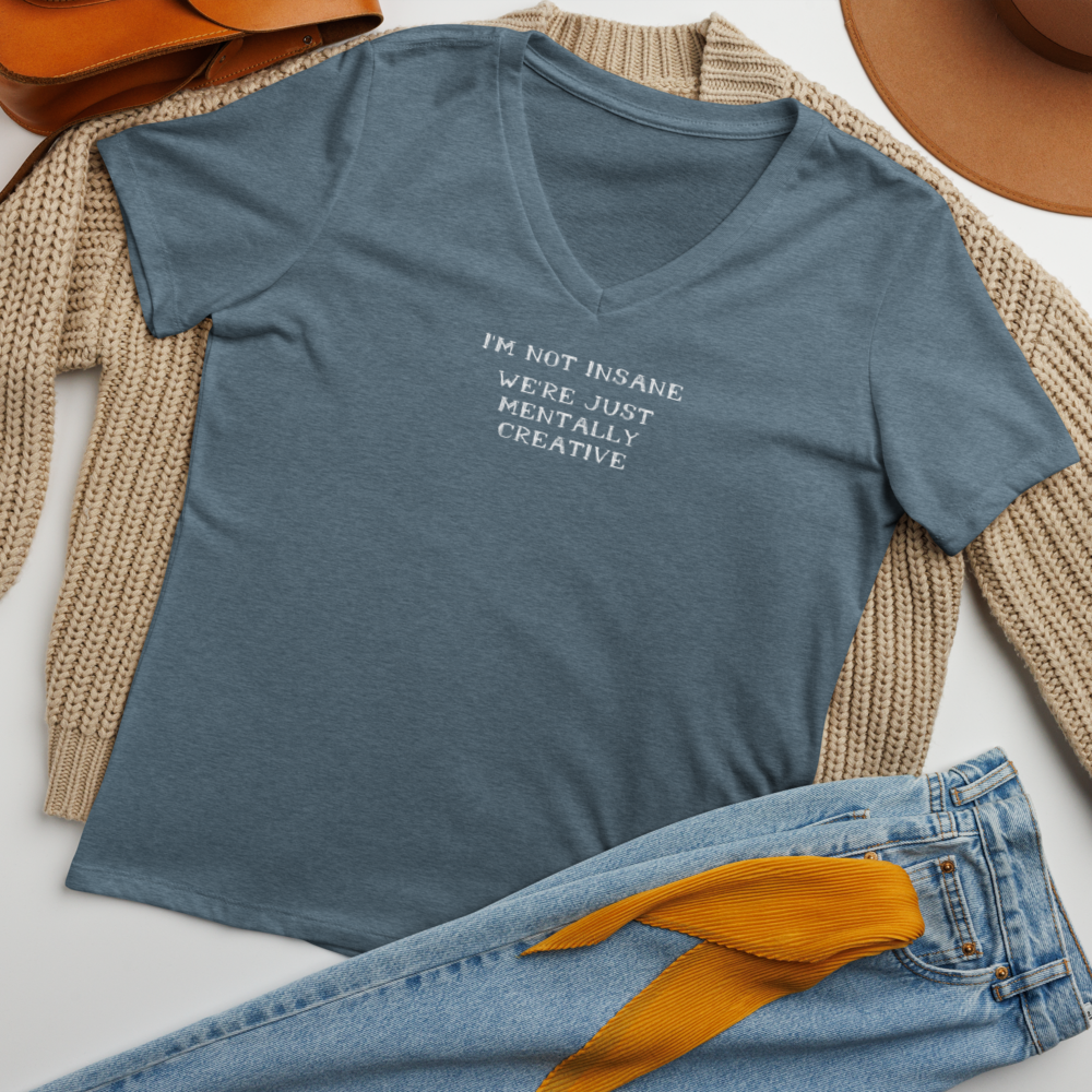 "I’m not insane   We’re just mentally creative" printed on a heather slate women's relaxed v-neck t-shirt laying on a flat surface amongst other clothes