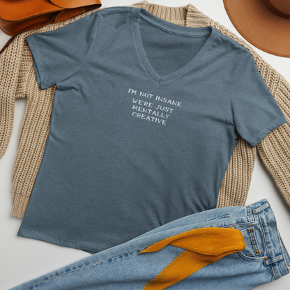 "I’m not insane   We’re just mentally creative" printed on a heather slate women's relaxed v-neck t-shirt laying on a flat surface amongst other clothes