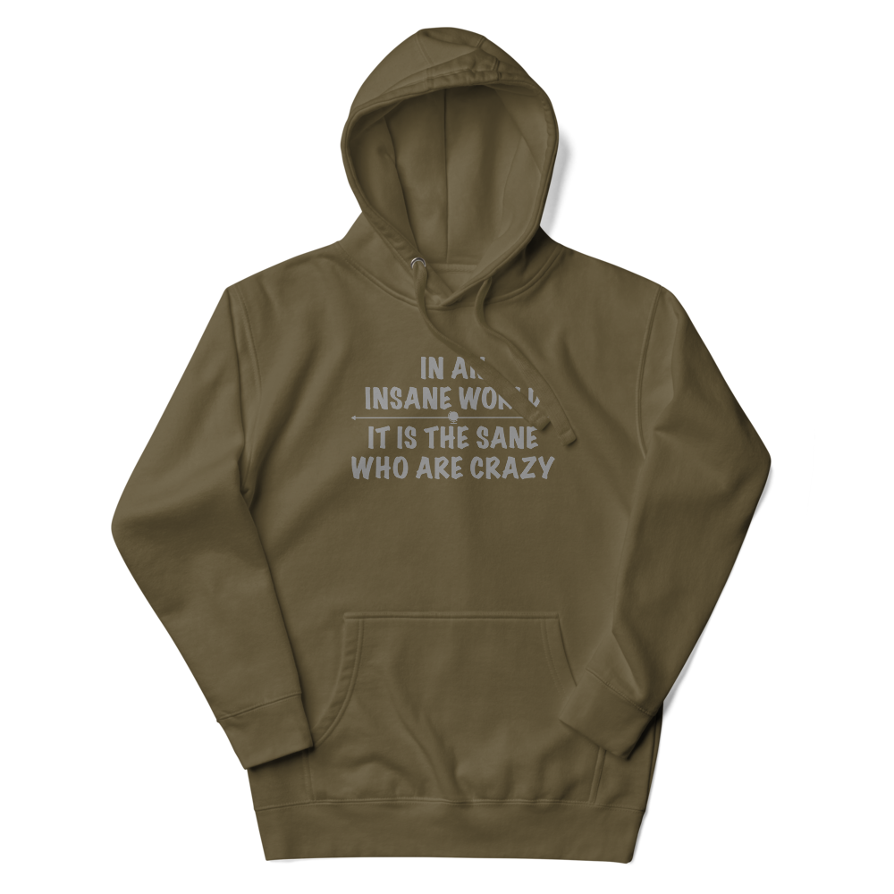 "In an insane world, it is the sane who are crazy" printed on a military green unisex hoodie laying on a flat surface with the hood up
