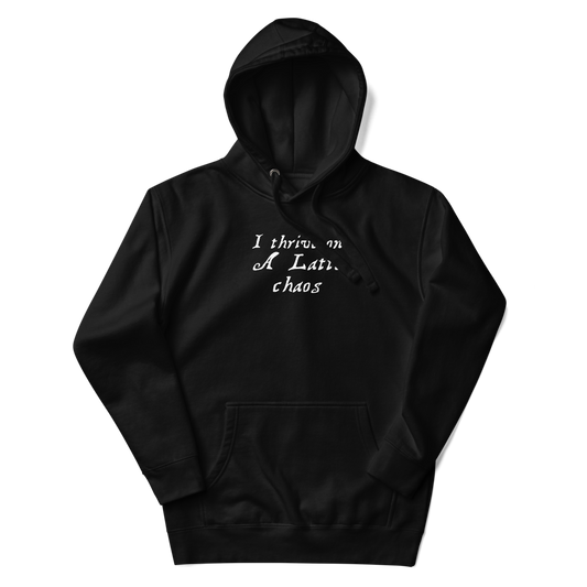 "I thrive on a latte chaos" printed on a black unisex hoodie laying on a flat surface with the hood up
