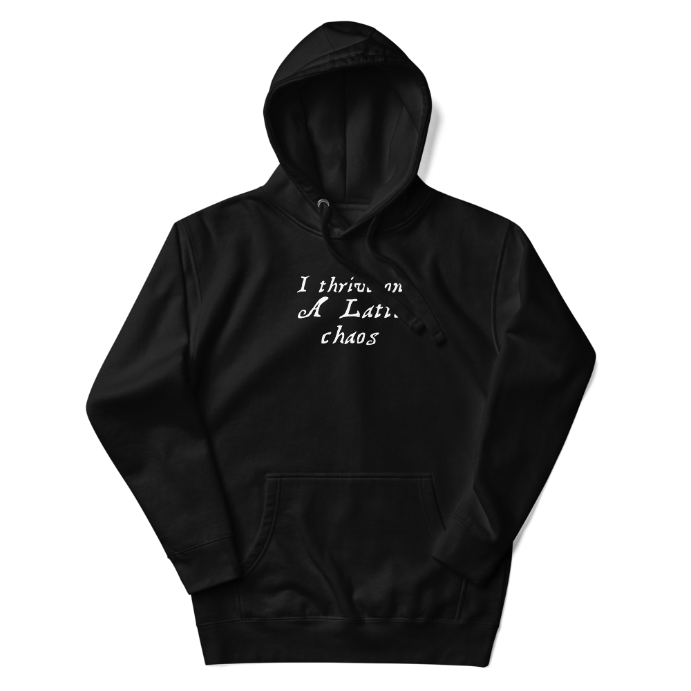"I thrive on a latte chaos" printed on a black unisex hoodie laying on a flat surface with the hood up