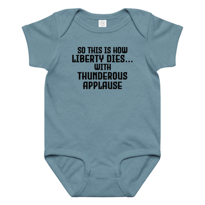 "So this is how liberty dies… with thunderous applause" printed on a vintage indigo baby jersey bodysuit laying on a flat surface amongst other clothes
