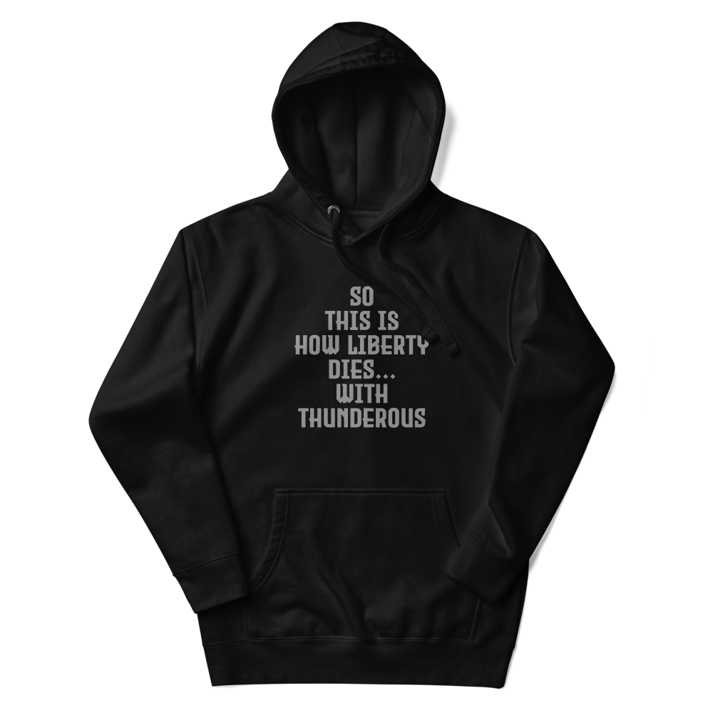 "So this is how liberty dies… with thunderous applause" printed on a black unisex hoodie laying on a flat surface with the hood up