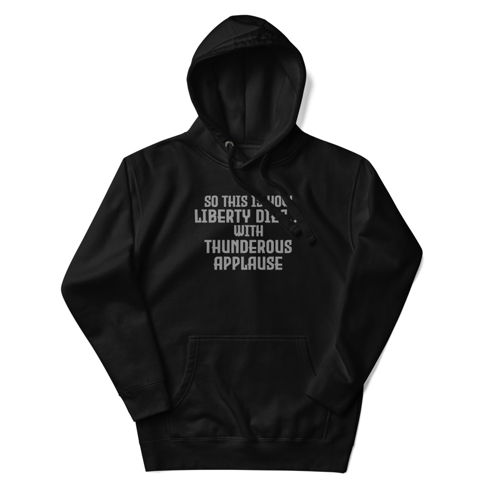 "So this is how liberty dies… with thunderous applause" printed on a black unisex hoodie laying on a flat surface with the hood up