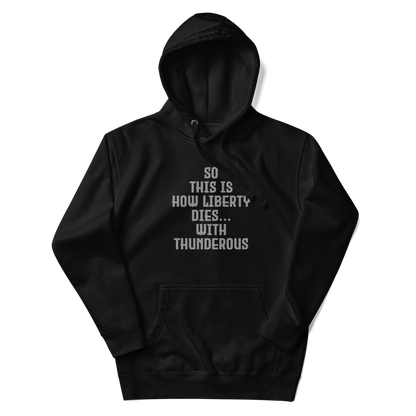 "So this is how liberty dies… with thunderous applause" printed on a black unisex hoodie laying on a flat surface with the hood up