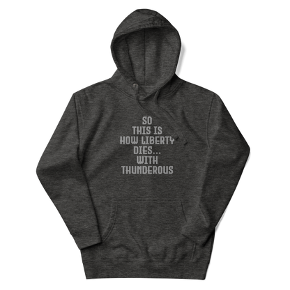 "So this is how liberty dies… with thunderous applause" printed on a charcoal heather unisex hoodie laying on a flat surface with the hood up