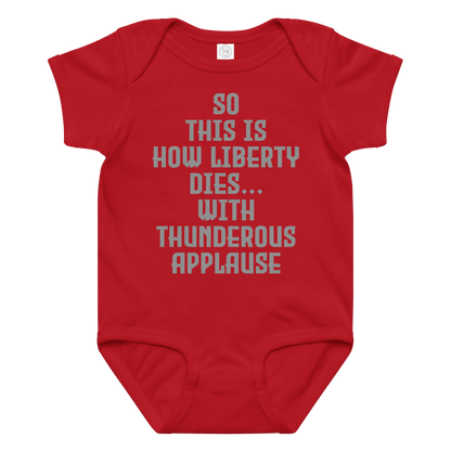 "So this is how liberty dies… with thunderous applause" printed on a red baby jersey bodysuit laying on a flat surface amongst other clothes