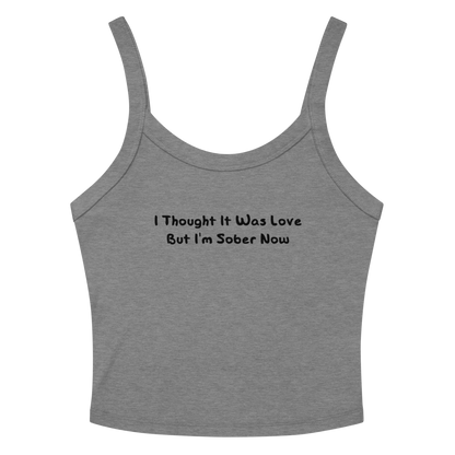 "I thought it was love but I’m sober now" printed on a athletic heather women's micro-rib tanktop laying on a flat surface amongst sunglasses and shorts