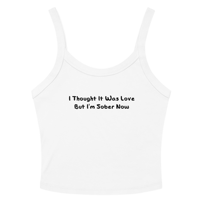 "I thought it was love but I’m sober now" printed on a solid white blend women's micro-rib tanktop laying on a flat surface amongst sunglasses and shorts