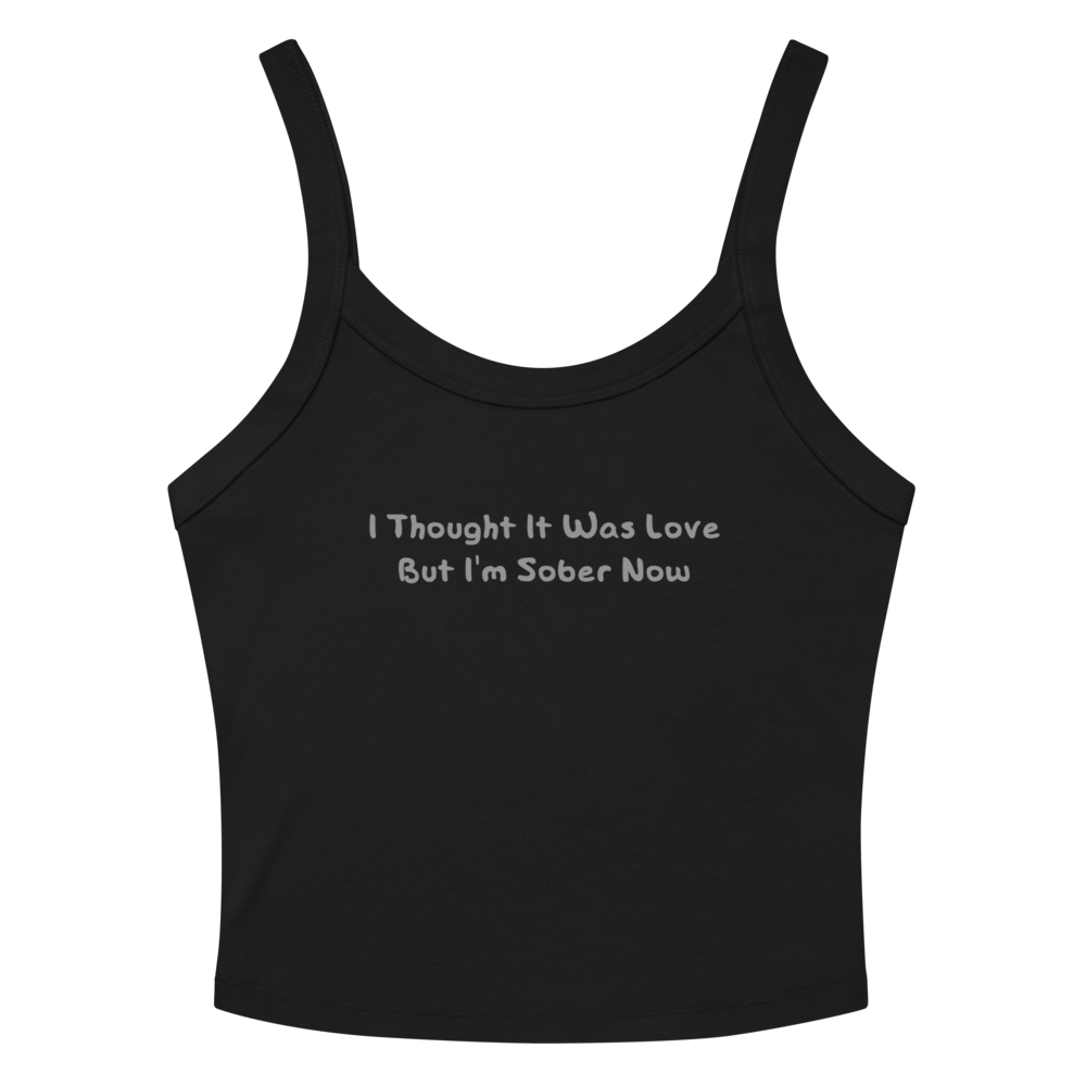 "I thought it was love but I’m sober now" printed on a solid black blend women's micro-rib tanktop laying on a flat surface amongst sunglasses and shorts