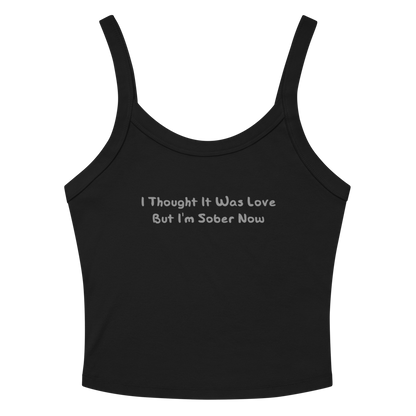 "I thought it was love but I’m sober now" printed on a solid black blend women's micro-rib tanktop laying on a flat surface amongst sunglasses and shorts