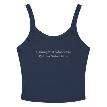 "I thought it was love but I’m sober now" printed on a solid navy blend women's micro-rib tanktop laying on a flat surface amongst sunglasses and shorts