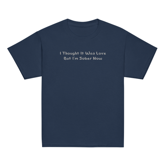 "I thought it was love but I’m sober now" printed on a navy youth classic tee laying on a flat surface