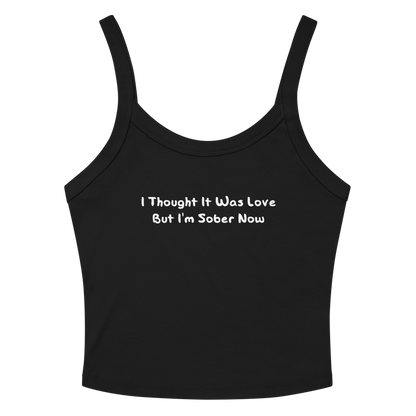 "I thought it was love but I’m sober now" printed on a solid black blend women's micro-rib tanktop laying on a flat surface amongst sunglasses and shorts