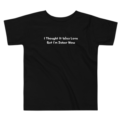 "I thought it was love but I’m sober now" printed on a black toddler short sleeve tee laying on a flat surface
