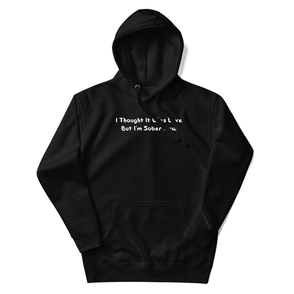 "I thought it was love but I’m sober now" printed on a black unisex hoodie laying on a flat surface with the hood up