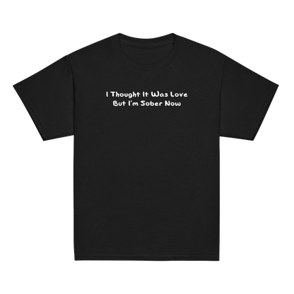 "I thought it was love but I’m sober now" printed on a black youth classic tee laying on a flat surface