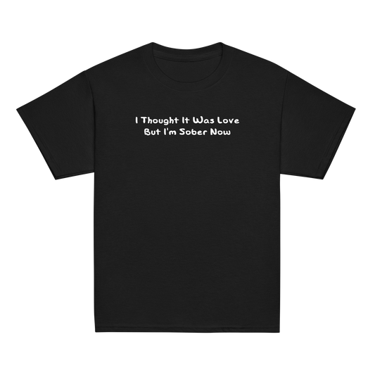 "I thought it was love but I’m sober now" printed on a black youth classic tee laying on a flat surface