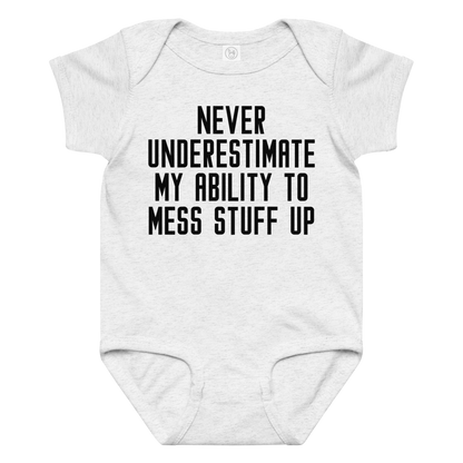 "Never Understimate My Ability To Mess Stuff Up" printed on a ash baby jersey bodysuit laying on a flat surface amongst other clothes