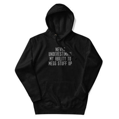"Never Understimate My Ability To Mess Stuff Up" printed on a black unisex hoodie laying on a flat surface with the hood up