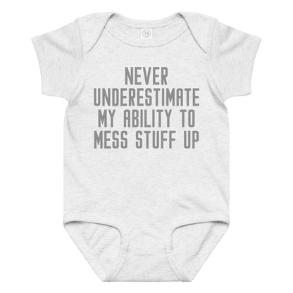 "Never Understimate My Ability To Mess Stuff Up" printed on a ash baby jersey bodysuit laying on a flat surface amongst other clothes