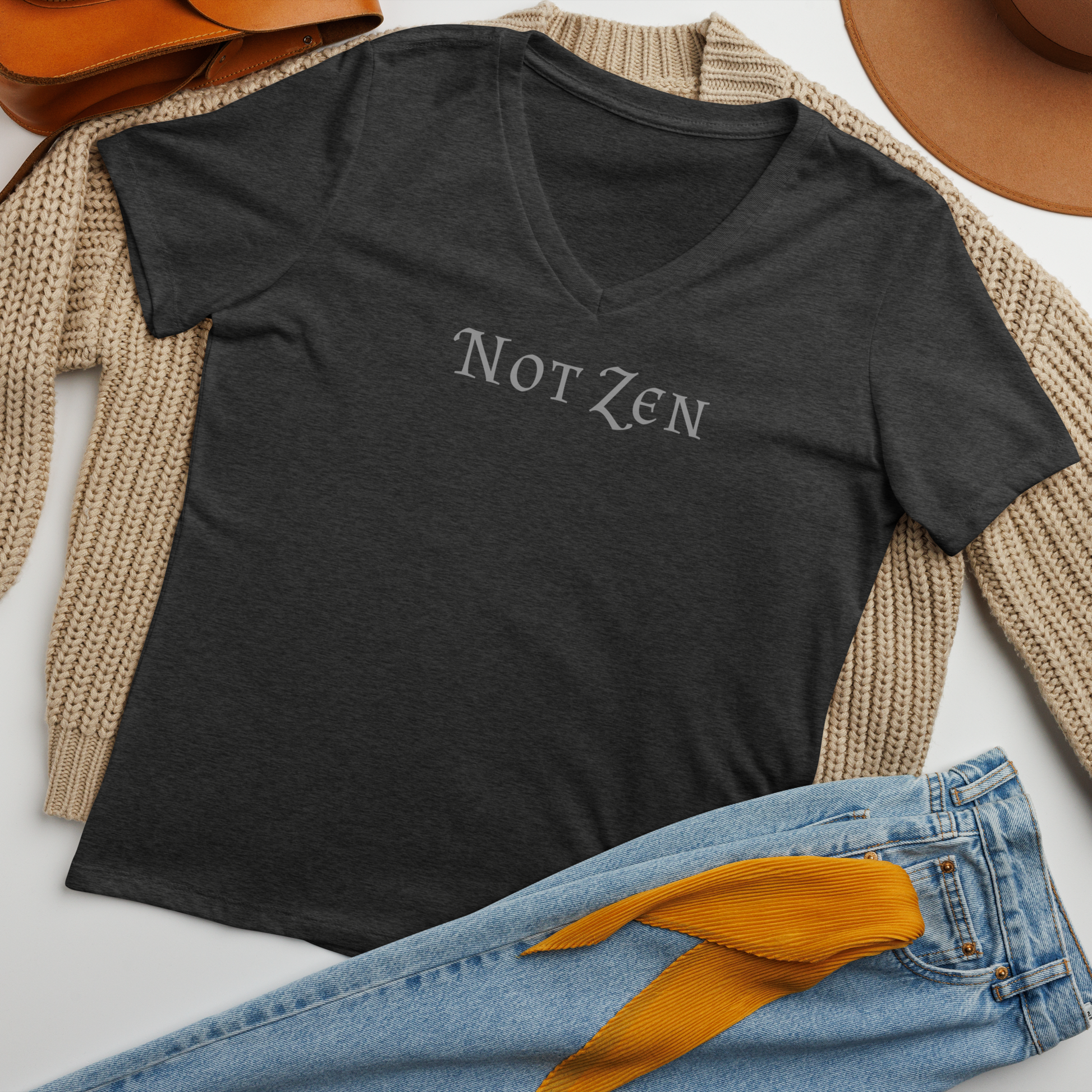 "Not zen" printed on a dark grey heather women's relaxed v-neck t-shirt laying on a flat surface amongst other clothes