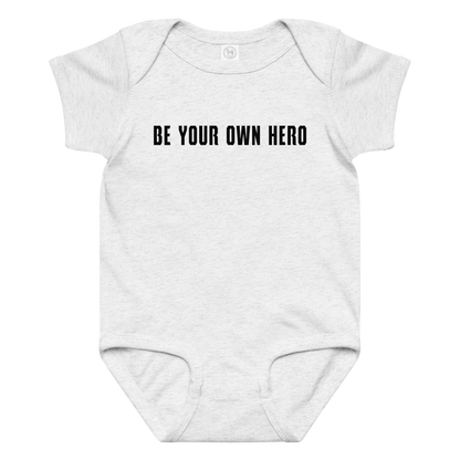 "Be your own here" printed on a ash baby jersey bodysuit laying on a flat surface amongst other clothes