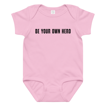 "Be your own here" printed on a pink baby jersey bodysuit laying on a flat surface amongst other clothes