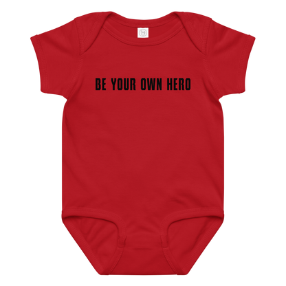 "Be your own here" printed on a red baby jersey bodysuit laying on a flat surface amongst other clothes
