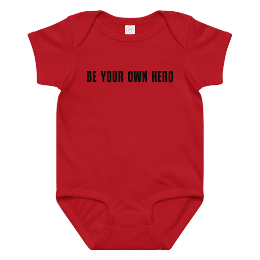 "Be your own here" printed on a red baby jersey bodysuit laying on a flat surface amongst other clothes