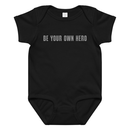 "Be your own here" printed on a black baby jersey bodysuit laying on a flat surface amongst other clothes
