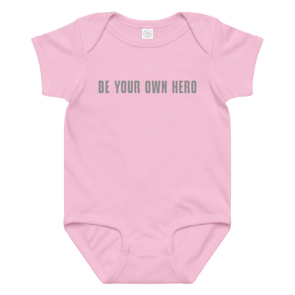 "Be your own here" printed on a pink baby jersey bodysuit laying on a flat surface amongst other clothes