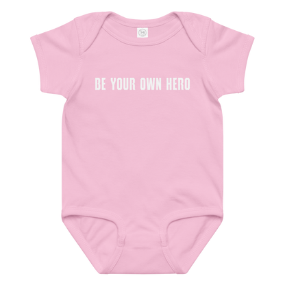 "Be your own here" printed on a pink baby jersey bodysuit laying on a flat surface amongst other clothes