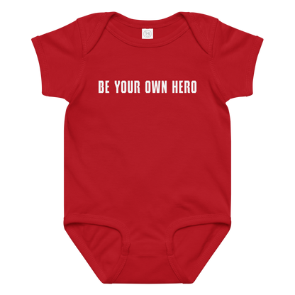 "Be your own here" printed on a red baby jersey bodysuit laying on a flat surface amongst other clothes
