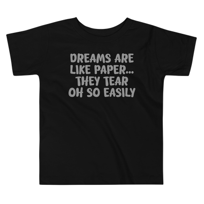 "Dreams are like paper, they tear oh so easily" printed on a black toddler short sleeve tee laying on a flat surface