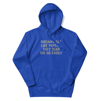 "Dreams are like paper, they tear oh so easily" printed on a team royal unisex hoodie laying on a flat surface with the hood up