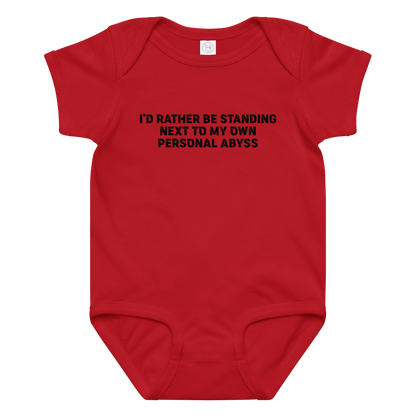 "I'd rather be standing next to my own personal abyss" printed on a red baby jersey bodysuit laying on a flat surface amongst other clothes