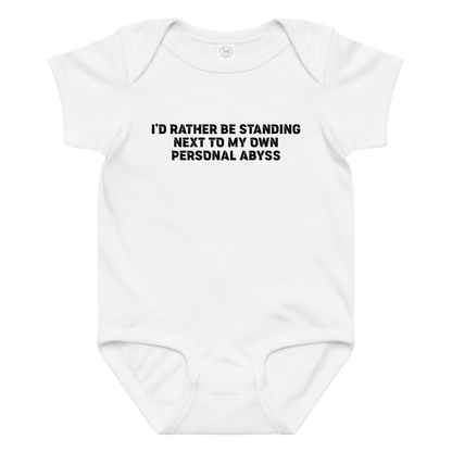 "I'd rather be standing next to my own personal abyss" printed on a white baby jersey bodysuit laying on a flat surface amongst other clothes