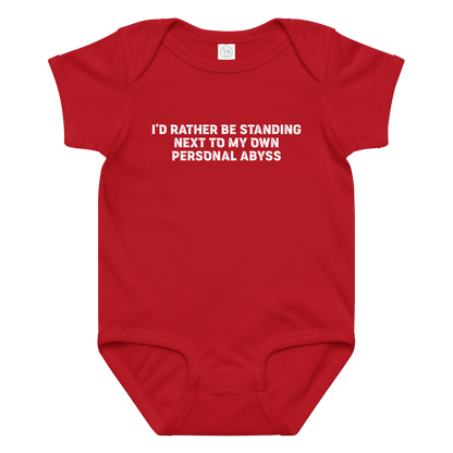 "I'd rather be standing next to my own personal abyss" printed on a red baby jersey bodysuit laying on a flat surface amongst other clothes