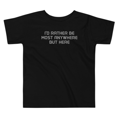 "I’d rather be most anywhere but here" printed on a black toddler short sleeve tee laying on a flat surface