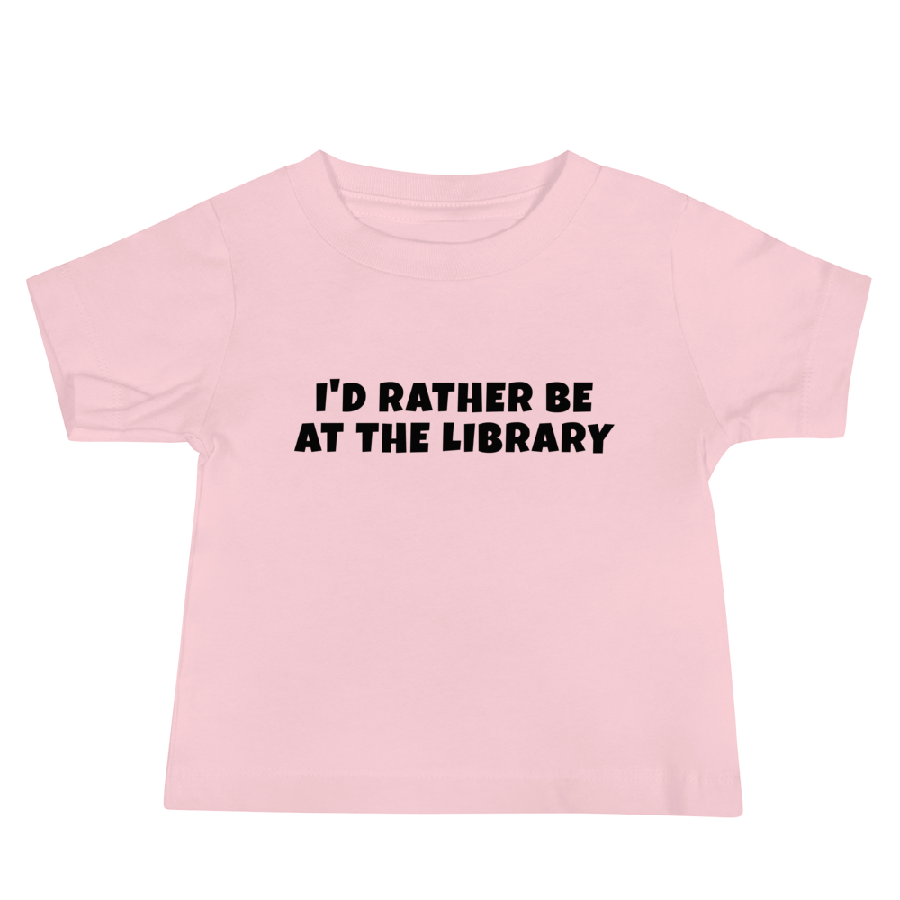 "I’d rather be at the library" printed on a pink baby jersey short sleeve tee laying on a flat surface