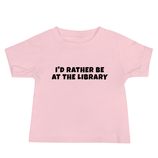 "I’d rather be at the library" printed on a pink baby jersey short sleeve tee laying on a flat surface