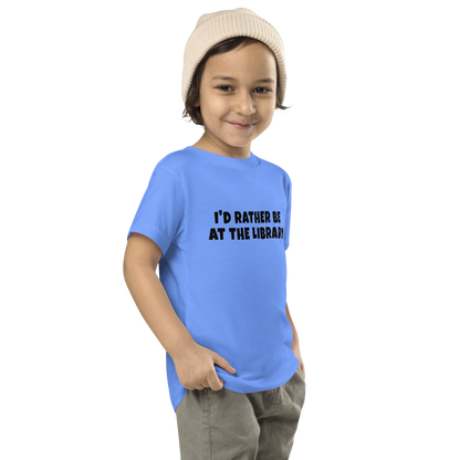 "I’d rather be at the library" printed on a heather columbia blue toddler short sleeve tee worn by a male who is also wearing a knit cap