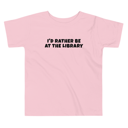 "I’d rather be at the library" printed on a pink toddler short sleeve tee laying on a flat surface
