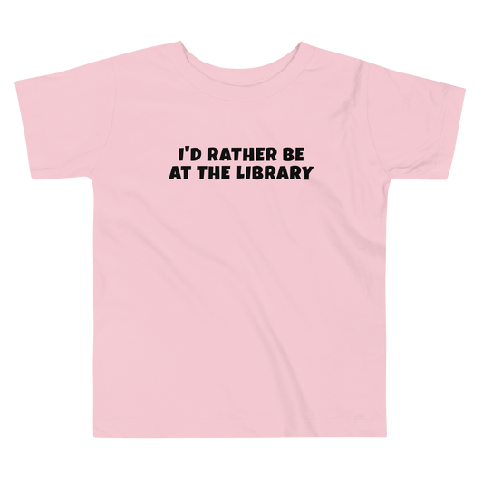 "I’d rather be at the library" printed on a pink toddler short sleeve tee laying on a flat surface