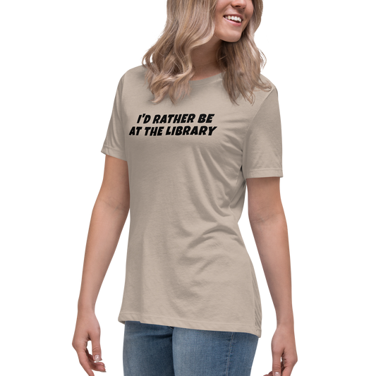 "I’d rather be at the library" printed on a heather stone women's relaxed t-shirt worn by a female