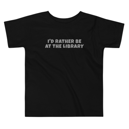 "I’d rather be at the library" printed on a black toddler short sleeve tee laying on a flat surface