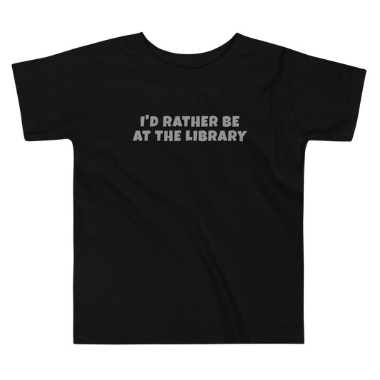 "I’d rather be at the library" printed on a black toddler short sleeve tee laying on a flat surface