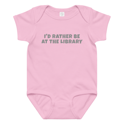 "I’d rather be at the library" printed on a pink baby jersey bodysuit laying on a flat surface amongst other clothes